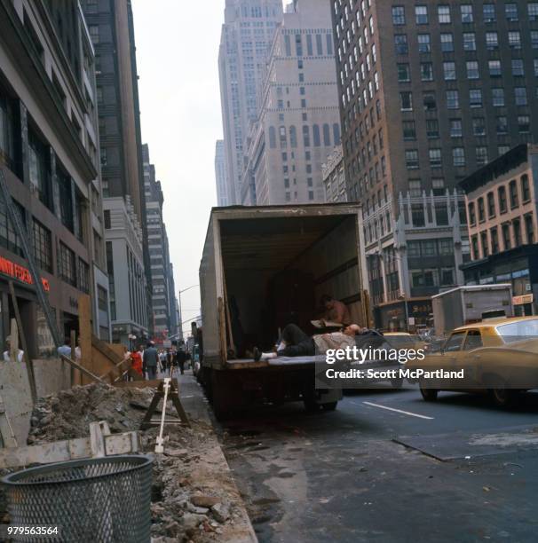 New York City - Beside a construction site, a man lies on the open tailgate of a truck; another man in the back of the truck reads a newspaper.