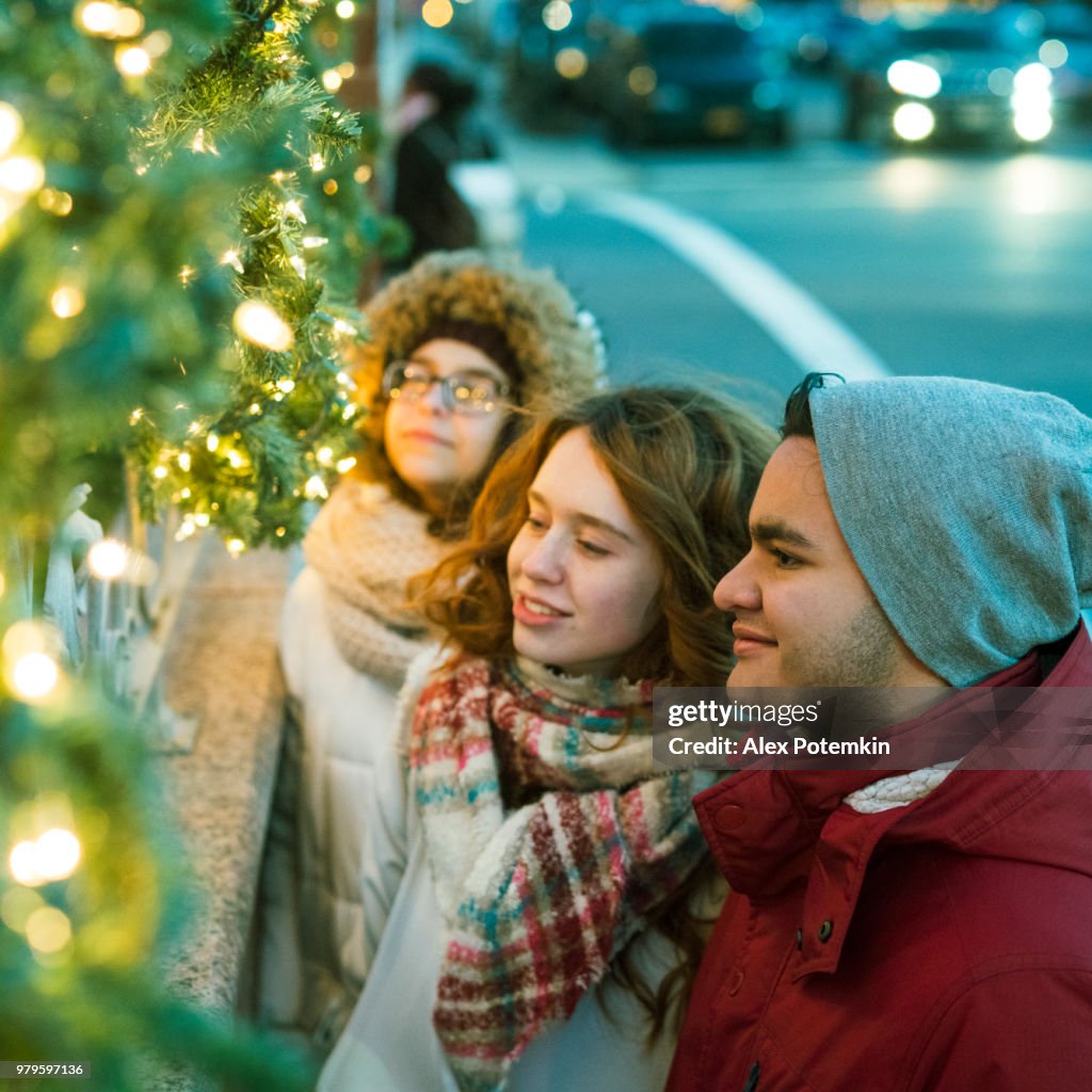 Interracial loving couple, teenagers, the beautiful Caucasian white 17-years-old long haired girl and handsome Latino Hispanic 18-years-old boy, and their friend, 15-years-old teenager girl, enjoying Christmas Illumination and having fun in the cold winte