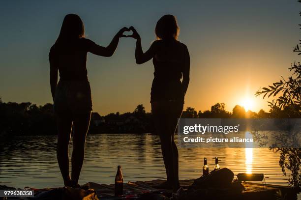 June 2018, Germany, Weissling: Lynn and Annika enjoy a lakeside picknick and form a heart with their hands as the sun sets. Photo: Lino Mirgeler/dpa