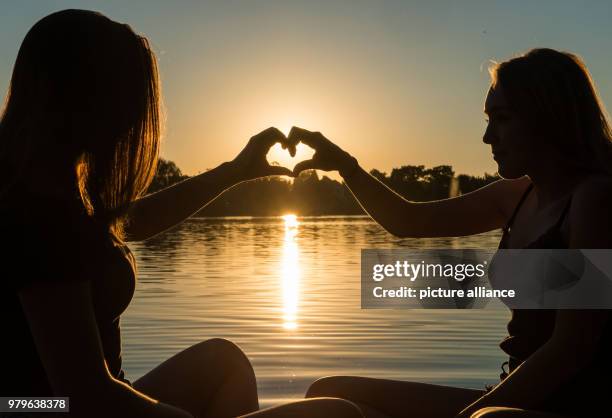 June 2018, Germany, Weissling: Lynn and Annika enjoy a lakeside picknick and form a heart with their hands as the sun sets. Photo: Lino Mirgeler/dpa