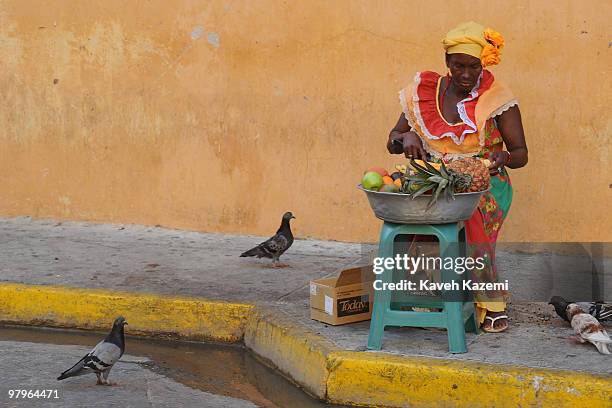 Of INDIAS, COLOMBIA A Cartagenan woman in a colorful traditional dress sells fresh fruits while sat on the street side, with pigeons sorrounding her,...