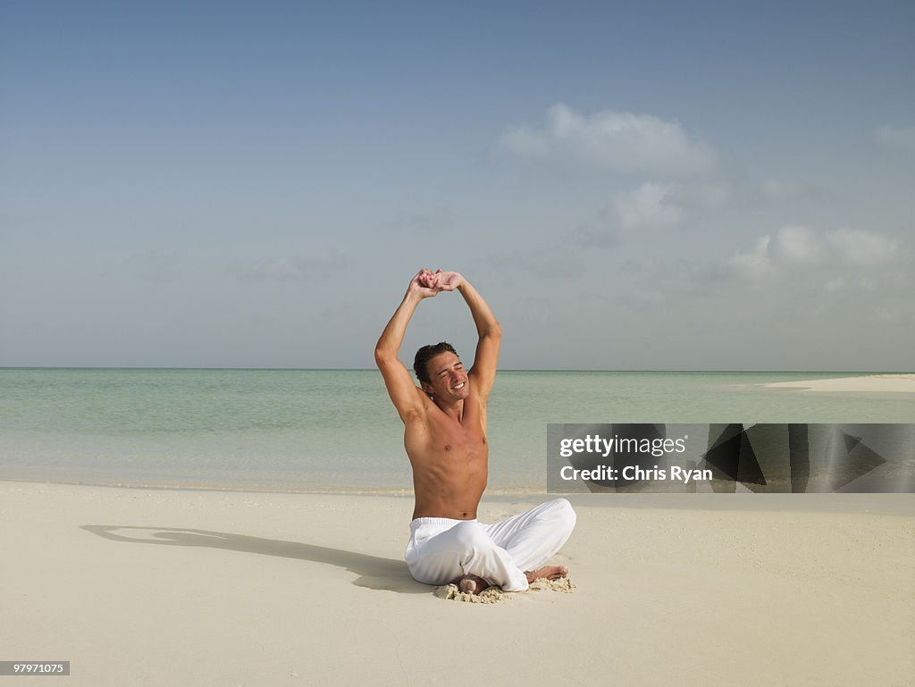 Man sitting cross-legged on beach and stretching arms overhead