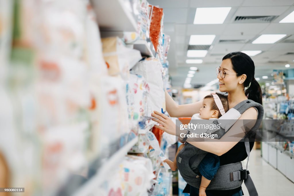 Young Asian mother carrying cute baby girl shopping for baby product in a shopping mall and is looking at a variety of diapers