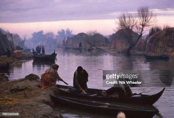 People and boat on river on edge of traditional Marsh Arab village of reed houses in the wetlands of Southern Iraq circa 1978.