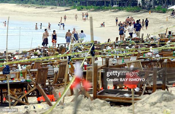 Tourists walk along a beach in the Jimbaran area of Bali island, 02 October 2005, as damaged chairs and tables of a restaurant are seen behind police...