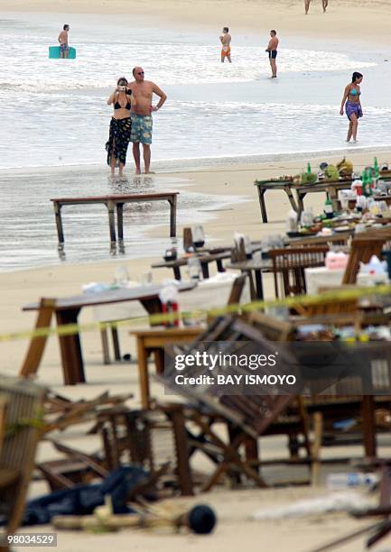 Tourists walk along a beach in the Jimbaran area of Bali island, 02 October 2005, as damaged chairs and tables of a restaurant are seen behind police...