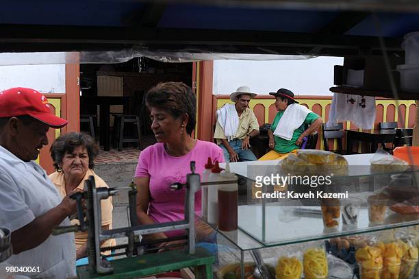 Street vendor sells fresh fruits while a typical Paisa man and a woman wearing traditional clothes and sombreros sit outside a bar on the main square...