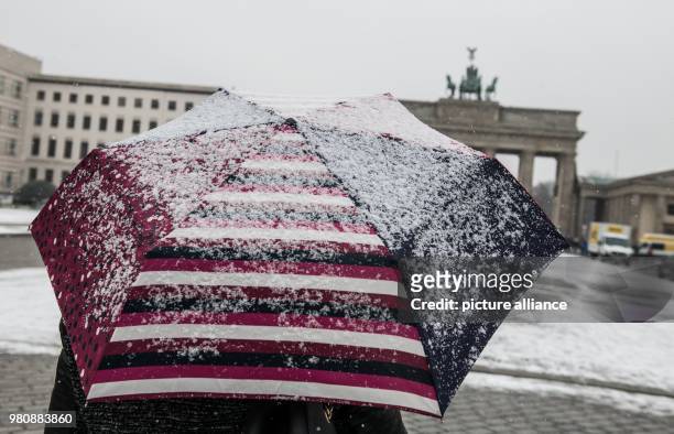 March 2018, Germany, Berlin: A woman standing during light snowfall with a snow-covered umbrella in front of the Brandenburg Gate. At the beginning...