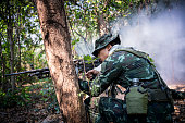 Special forces soldier  or private millitary holding gun aiming behind a tree at field and smoke background.