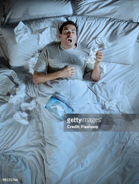 man lying in bed sneezing into a tissue - illness stock pictures, royalty-free photos & images