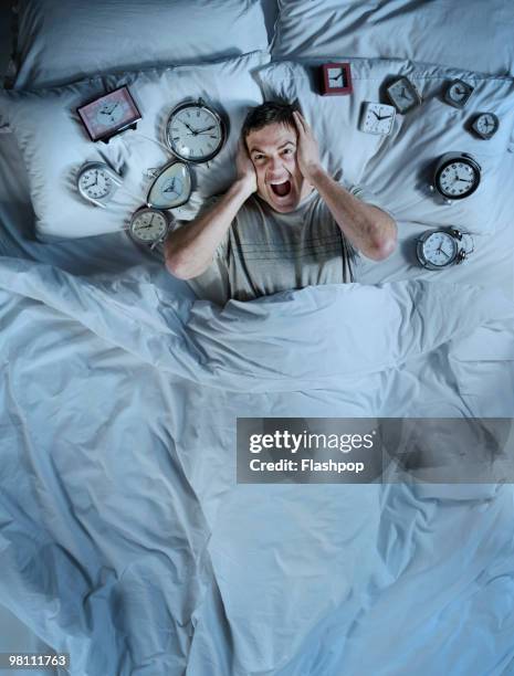 man screaming in bed surrounded by alarm clocks  - insonnia foto e immagini stock