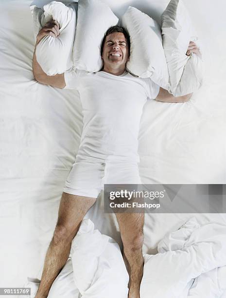 man in bed using pillows to cover ears - supino foto e immagini stock