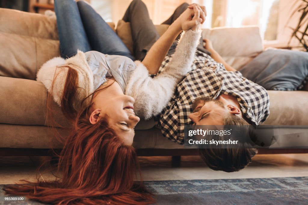 Coppia giocosa che si diverte mentre si tiene per mano e parla a testa in giù sul divano.