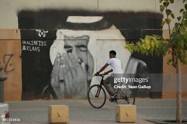 Man on a bicycle rides past a street artist's rendition of Saudi King Salman bin Abdulaziz praying on June 20, 2018 in Riyadh, Saudi Arabia. The...