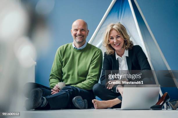 happy businessman and businesswoman sitting at teepee indoors with laptop - anti ageing stockfoto's en -beelden