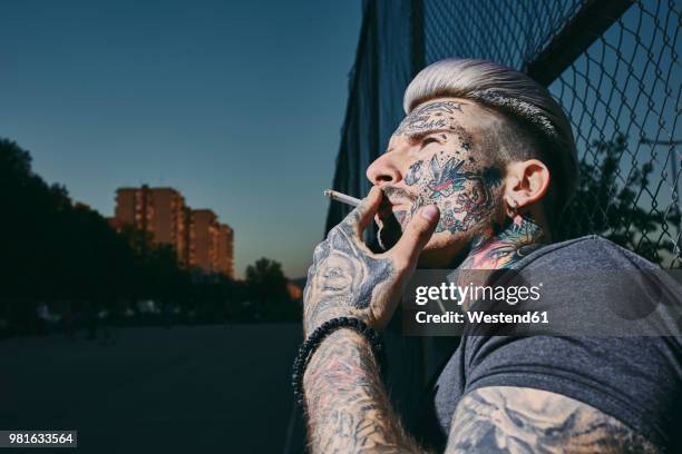 portrait of tattooed young man smoking a cigarette at wire mesh fence - anti smoking bildbanksfoton och bilder