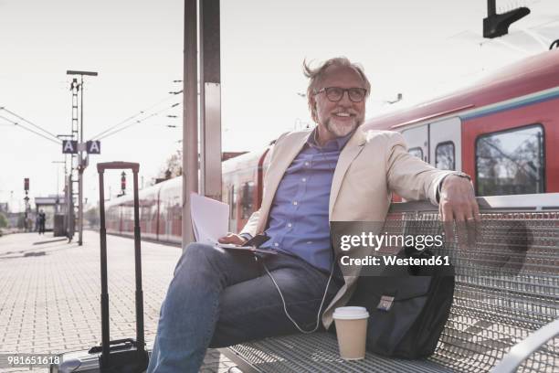 smiling mature businessman sitting at train station with cell phone, earbuds and notebook - bahnreisender stock-fotos und bilder