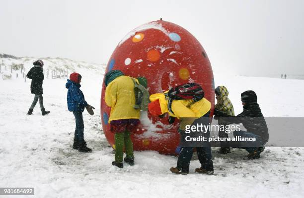 April 2018, Germany, Hiddensee Vitte: Visitors contemplate a snow-covered giant egg on the beach. The winter has struck once again short before...