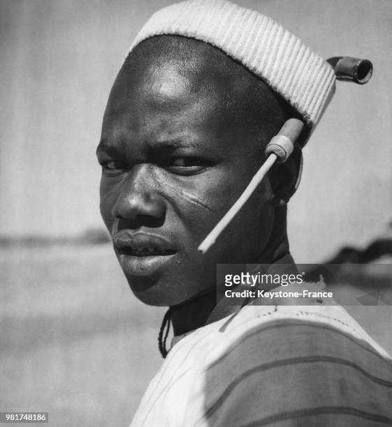 Jeune homme de la tribu Kouroumba, la tête rasée, coiffé d'une couronne de paille recouverte de coton et dans laquelle il glisse sa pipe.