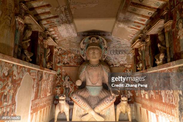 Buddha statue in the cave, surrounded with ancient colorful murals. The Mogao Caves, also known as the Thousand Buddha Grottoes, are the best known...