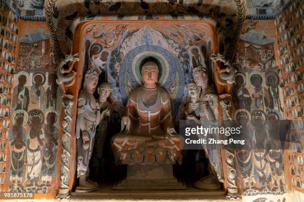 Buddha statue in the cave, surrounded with ancient colorful murals. The Mogao Caves, also known as the Thousand Buddha Grottoes, are the best known...