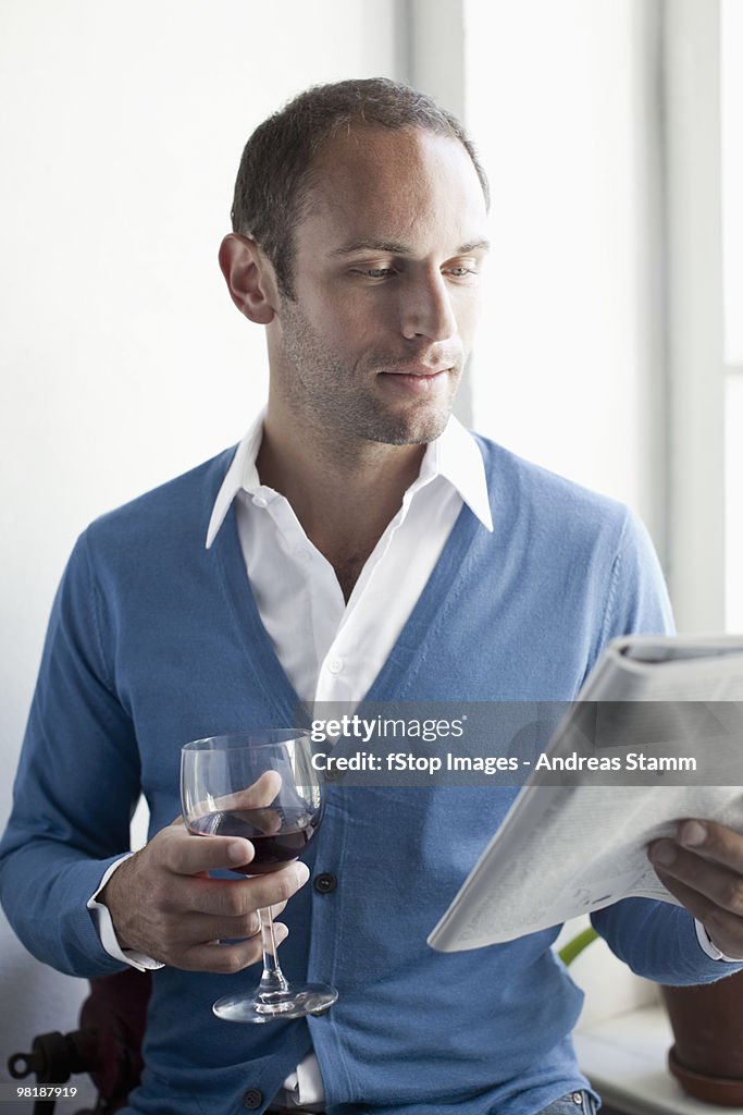 A man drinking a glass of wine and reading a magazine