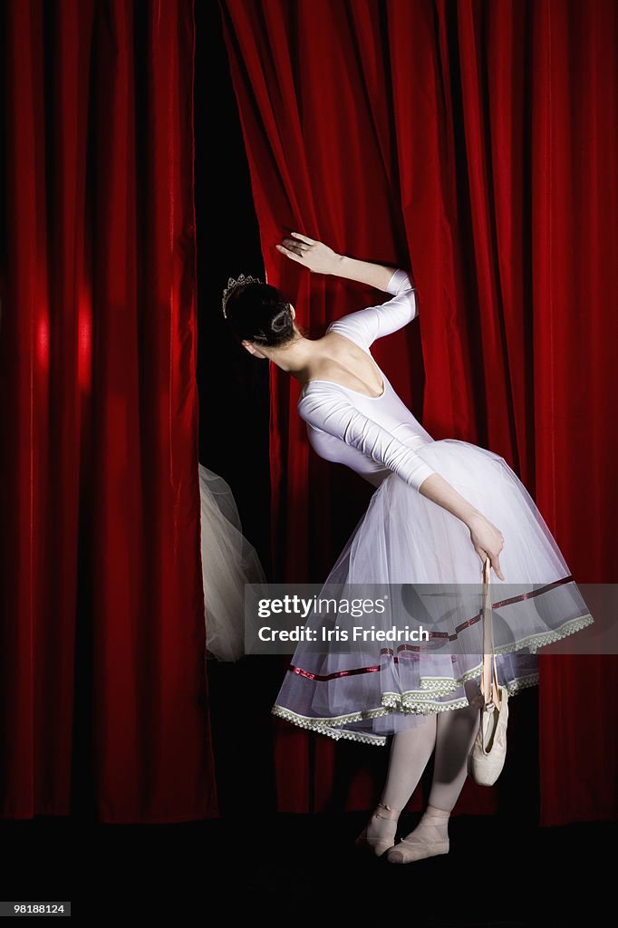 A ballet dancer peeking through a stage curtain holding a pointe shoe, rear view