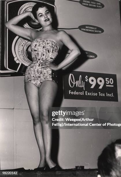 Model poses in front of a DeJUR camera, New York, ca.1950s. Behind her, a sign reads "Only $99.50, Federal Excise Tax Incl.".