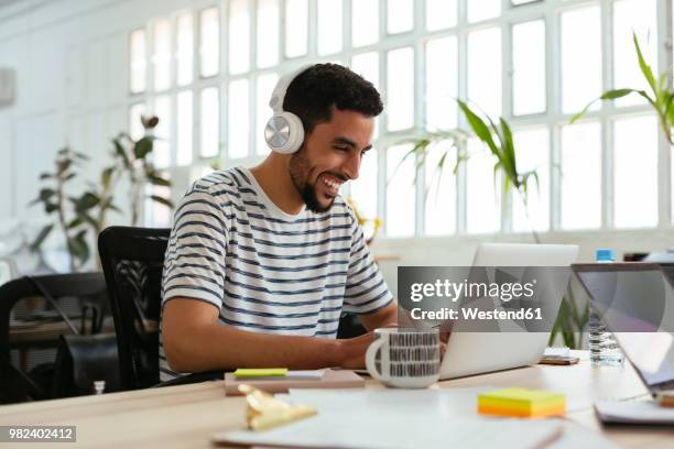 laughing young man wearing headphones using laptop at desk in office - black headphones stock pictures, royalty-free photos & images