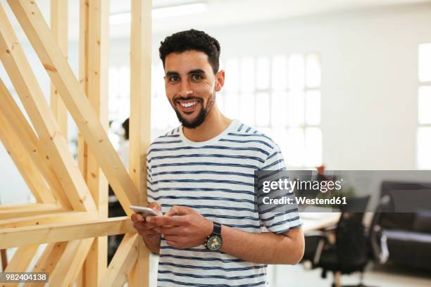 portrait of smiling young man with cell phone in office - nordafrikanischer abstammung stock-fotos und bilder