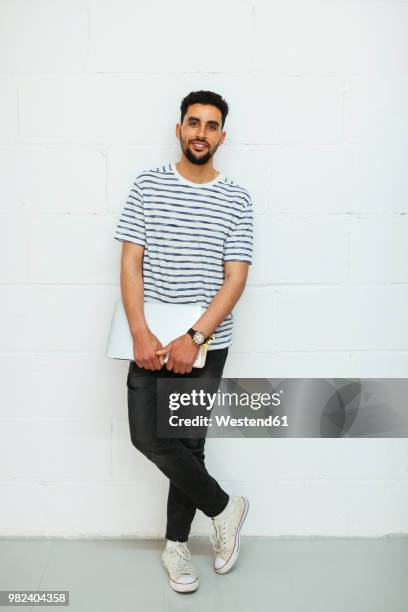 portrait of smiling young man with laptop standing at brick wall - holding laptop stock pictures, royalty-free photos & images