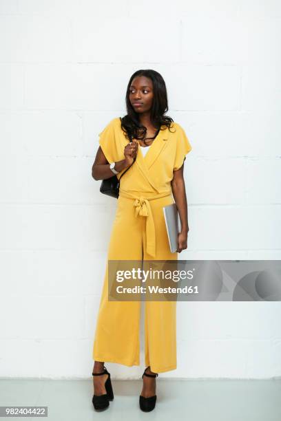 portrait of young woman with laptop standing at brick wall - jumpsuit stock-fotos und bilder