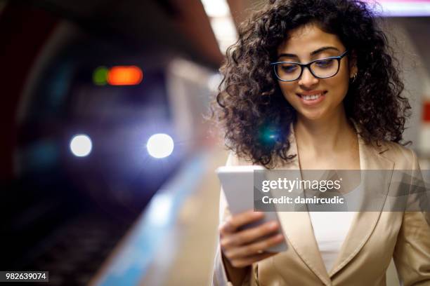 jovem empresária de pé na estação de metrô e usando o celular enquanto aguarda o trem - damircudic - fotografias e filmes do acervo