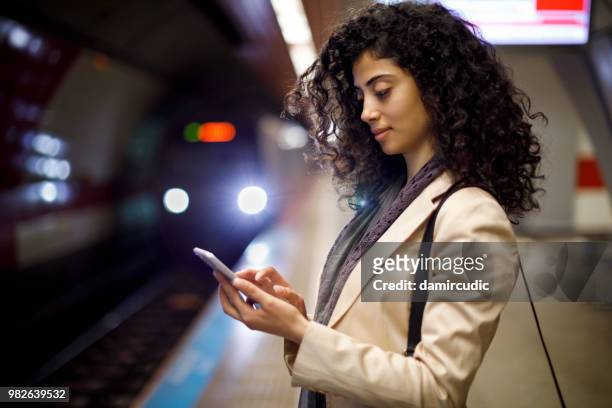 young businesswoman standing on the subway station and using her mobile phone while waiting for the train - damircudic stock pictures, royalty-free photos & images