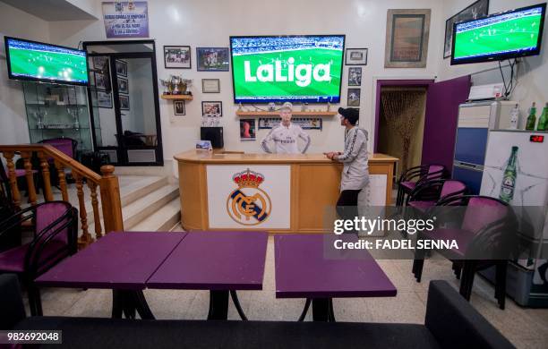 Supporter of Real Madrid football team watches a match on a TV screen above a wooden cut-out of Cristiano Ronaldo lying behind a reception counter,...