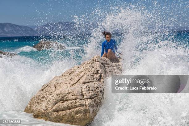 young woman sitting on rock hit by sea waves, cala gonone, sardinia, italy - febbraio stock-fotos und bilder