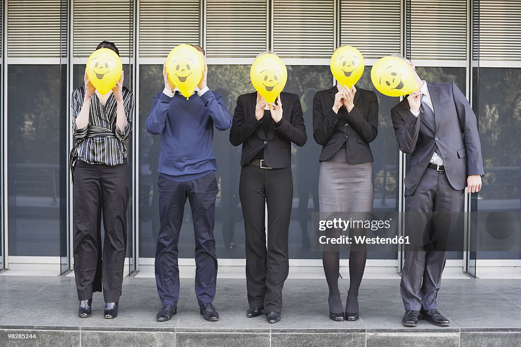 Germany, Hamburg, Five Business people standing in front of office building, hiding faces behind balloons