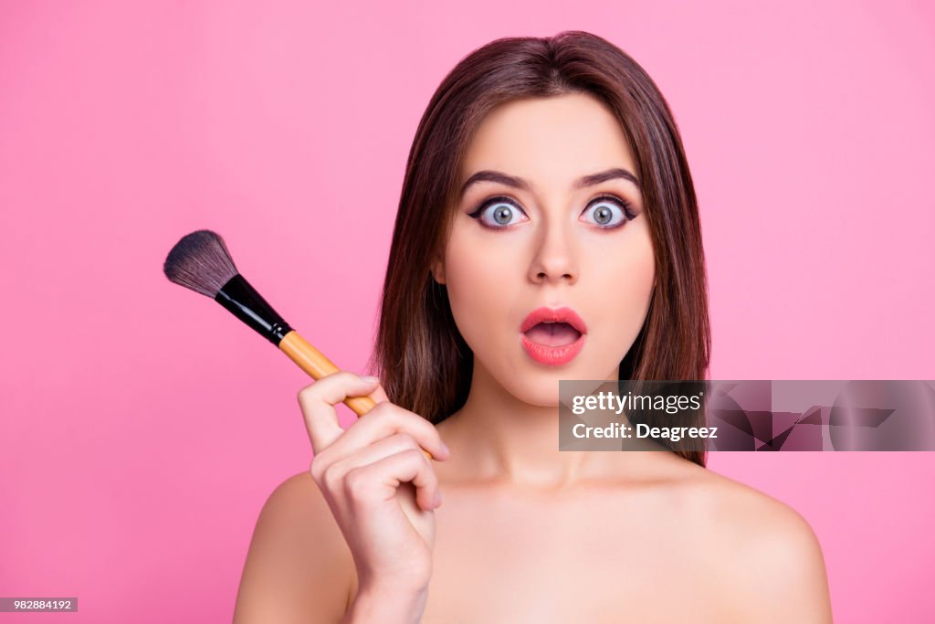 Close up portrait of shocked beautiful attractive pop-eyed pretty surprised scared make up artist holding a brush for blusher and powder in a hand, isolated on pink background
