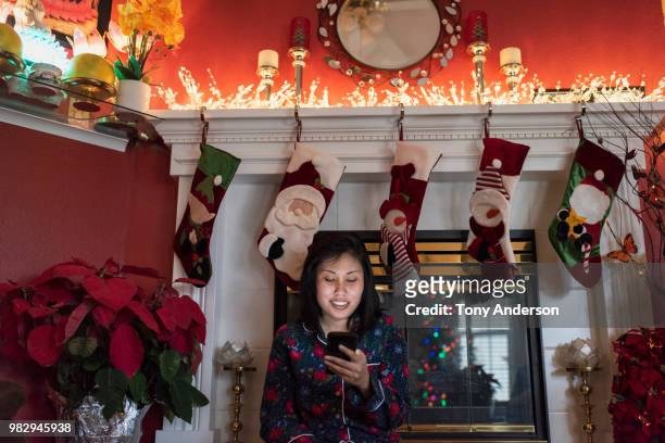 young woman with phone sitting on hearth decorated for christmas - stocking tops stock-fotos und bilder