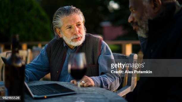 zwei alte männer, kaukasische weiß und schwarz, diskutieren investitionen mit laptop und trinken den wein im weingut - alex potemkin or krakozawr stock-fotos und bilder