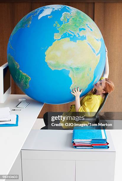 businesswoman looking up at large globe on lap in office - big tom stock-fotos und bilder