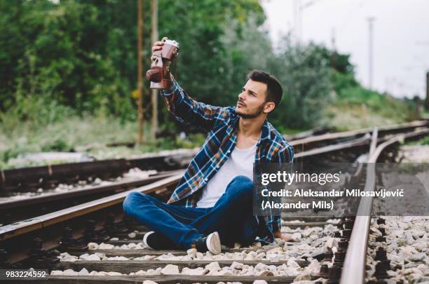 young hipster guy tourist with camera and bag taking a selfie on the railroad - camera bag stock-fotos und bilder