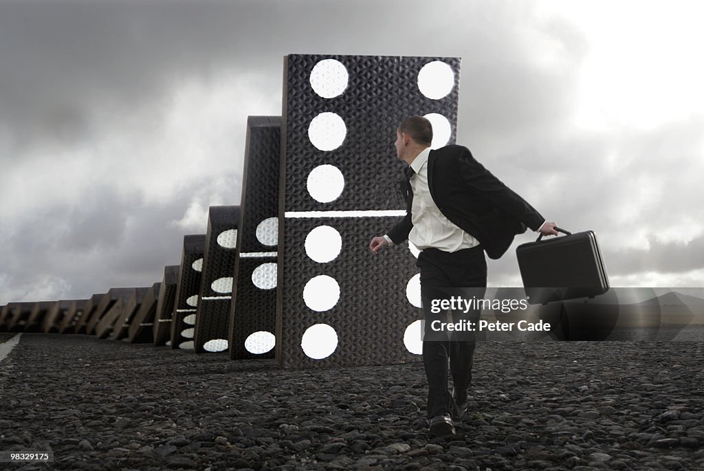 Male executive running from giant falling dominoes