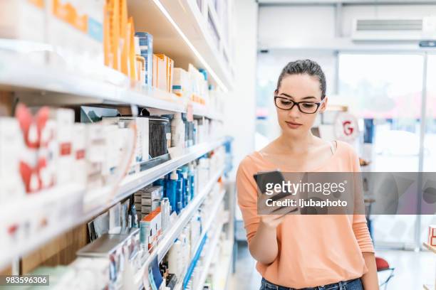 woman uses smart phone while in pharmacy - pharmacy imagens e fotografias de stock