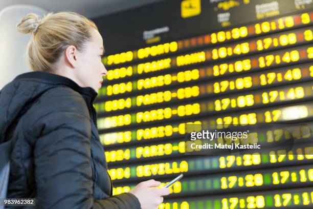 woman at airport in front of flight information board checking her phone. - departure board front on stockfoto's en -beelden