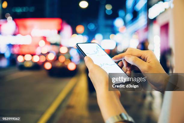 close up of woman's hand checking financial investment data on mobile phone in downtown city street at night - nasdaq building stock pictures, royalty-free photos & images