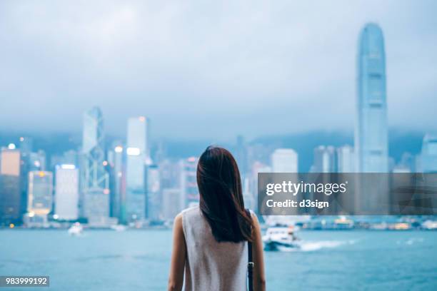 rear view of female traveller enjoys the view at victoria harbour and hong kong city skyline at dusk - über etwas schauen stock-fotos und bilder