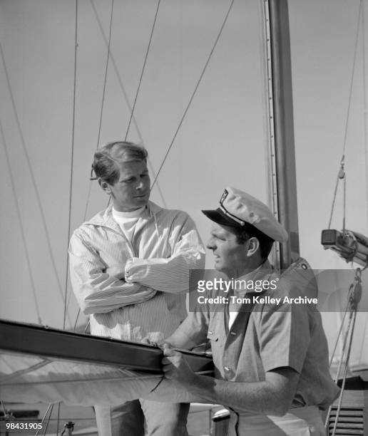 Man in a captain's cap and a shortsleeved shirt makes an adjustment to the boom of a sailboat as he talks with a man in a windbreaker, who listens...