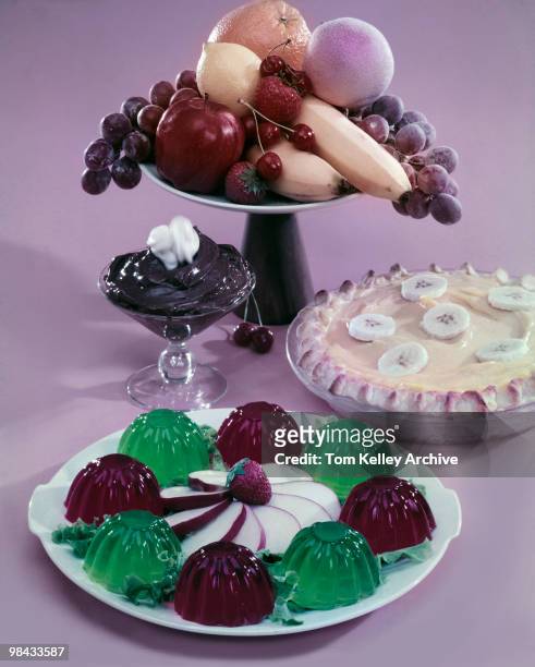 View of several dessert offerings, including molded red & green gelatin garnished with sliced apple, a banana-cream pie, chocolate pudding, and an...