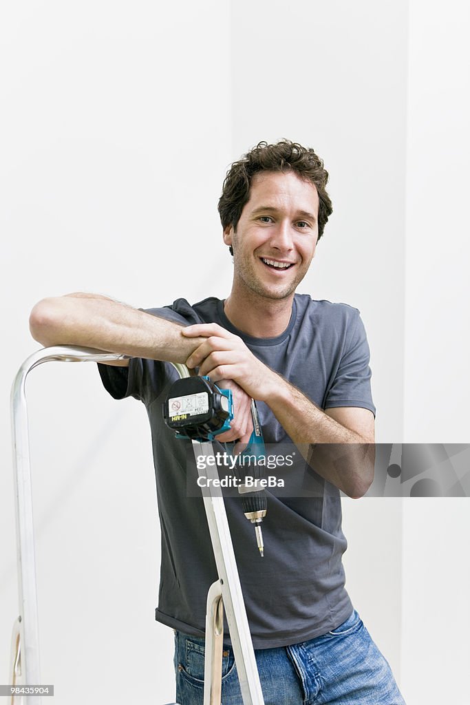 Portrait of young man with cordless screwdriver leaning against ladder